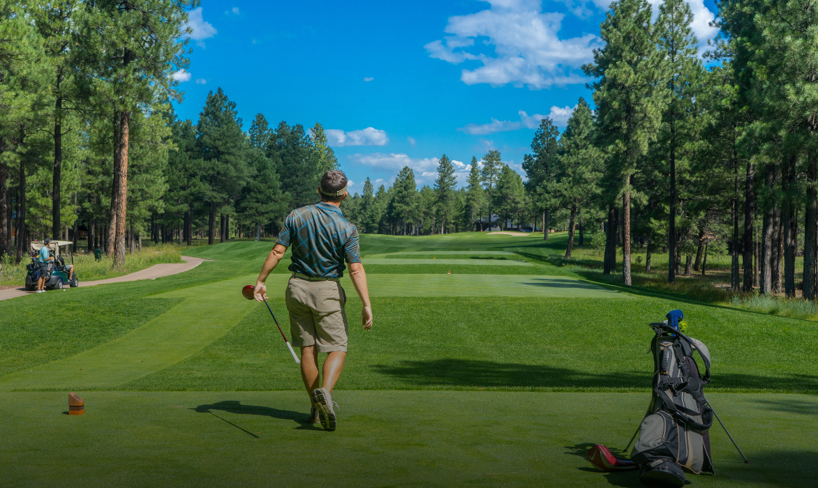Golfer in a polo shirt walking off a pine-lined tee, his bag on its stand